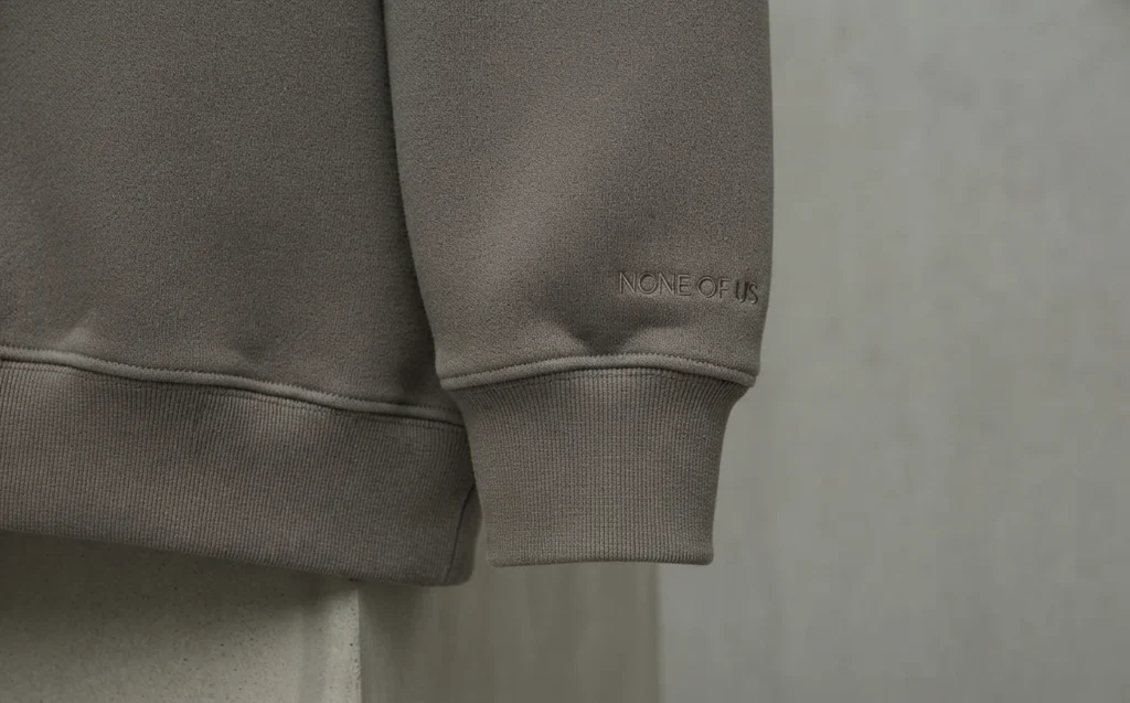 Close-up of the precision stitching, ribbed cuff, and hem of the charcoal grey None of Us (nofs) hoodie, emphasizing craftsmanship.