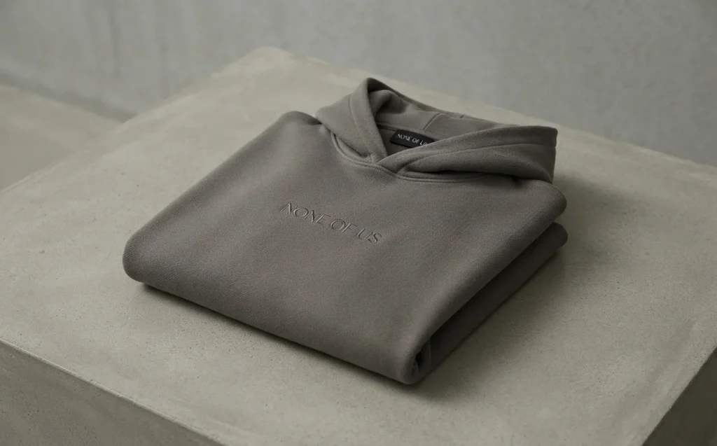 Charcoal grey heavyweight None of Us (nofs) branded hoodie neatly folded on a concrete surface, showcasing fabric texture.